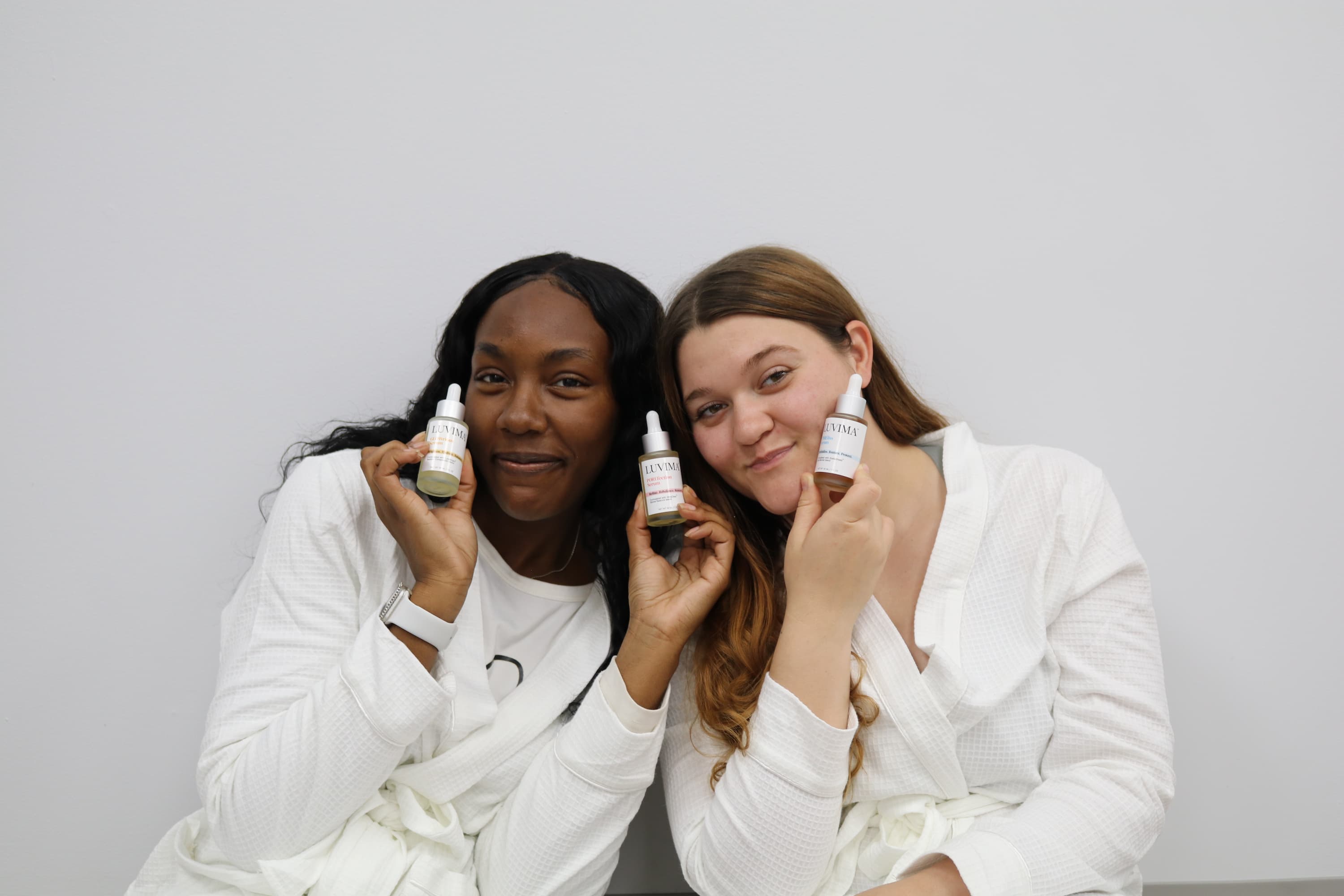 Two women smiling, wearing white robes, holding three LUVIMA serum bottles next to their faces against a gray background.