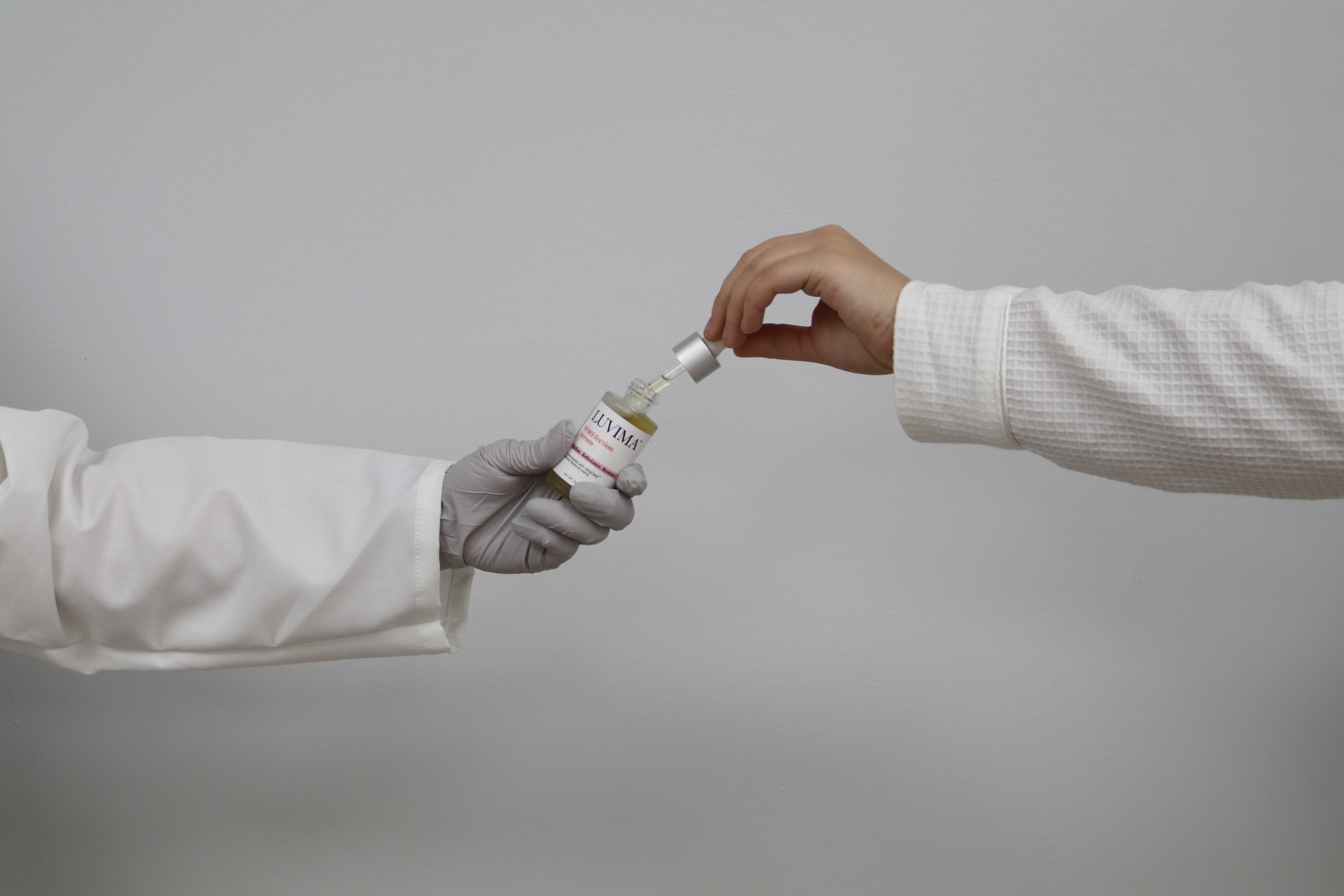 Two hands, one wearing a glove, reaching towards each other with a dropper bottle on a plain background